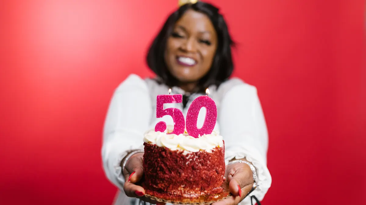 Femme joyeuse en chemise à manches longues blanches tenant un gâteau d'anniversaire avec bougies 50 sur fond rouge.