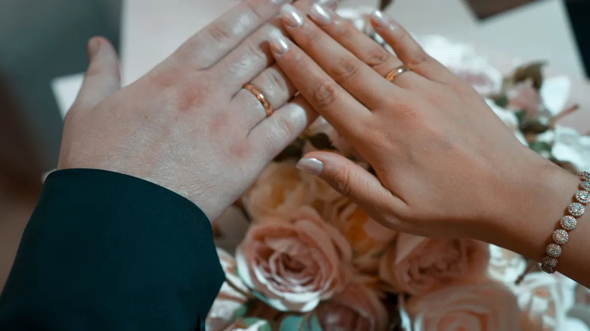 Gros plan sur des mains portant des alliances au-dessus d'un bouquet de roses roses.