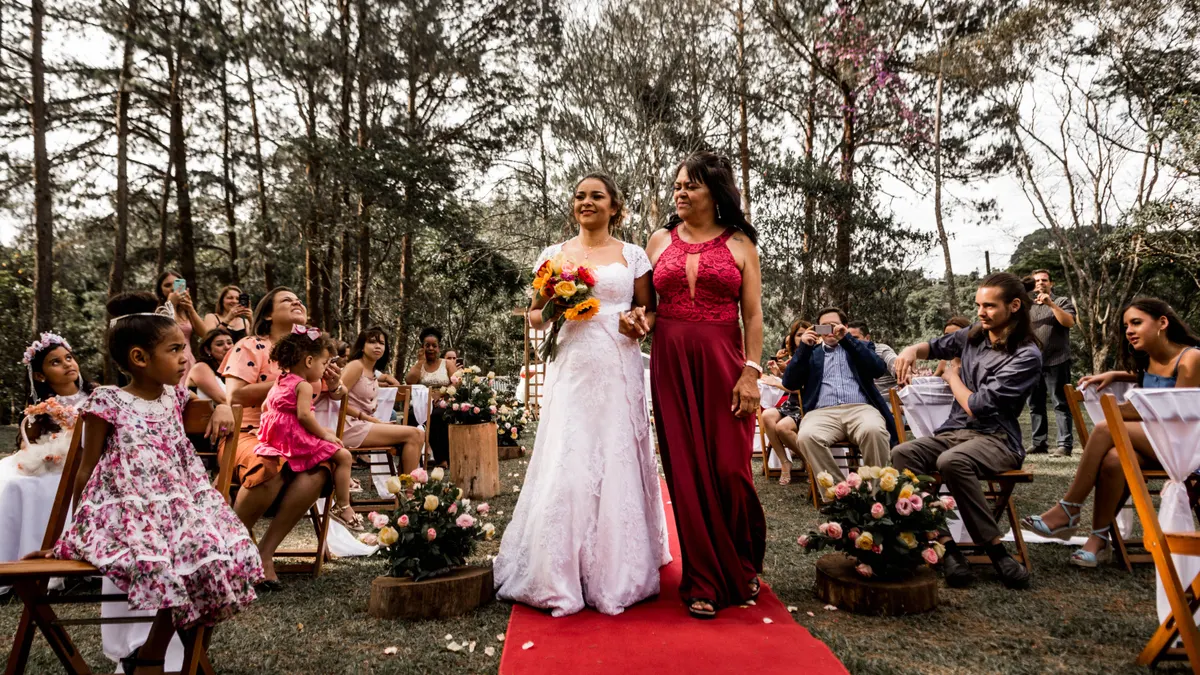 Mariée joyeuse marchant avec sa mère sur un tapis rouge à une cérémonie de mariage en plein air entourée d'invités.