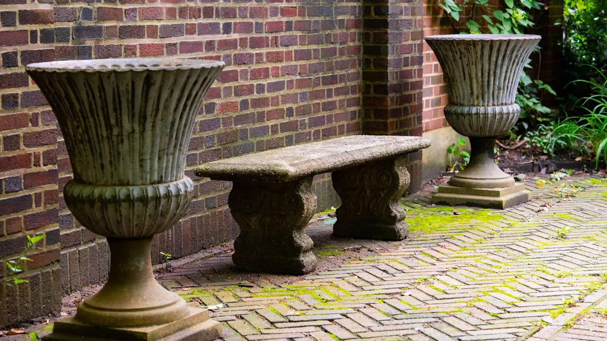 Banc en pierre classique et urnes dans un coin de jardin paisible avec allée en briques.